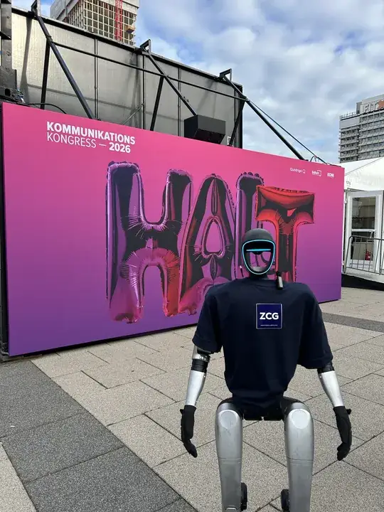 Humanoider Roboter im Shirt mit ZCG-Logo vor der Wand des Kommunikationskongresses 2026