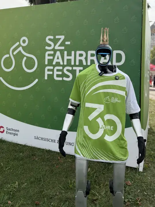 Humanoider Roboter im grünen Jubiläumstrikot des SZ-Fahrradfests vor der Sponsorenwand