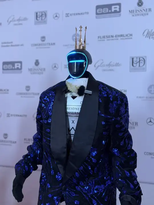 Humanoider Roboter in Nahaufnahme mit goldener Krone und blauem Pailletten-Sakko vor der Sponsorenwand